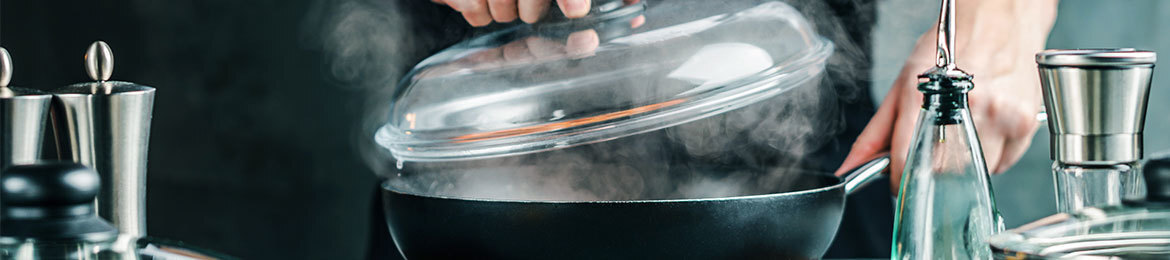 A cook lifts the glass lid from a pan, steam flows out.