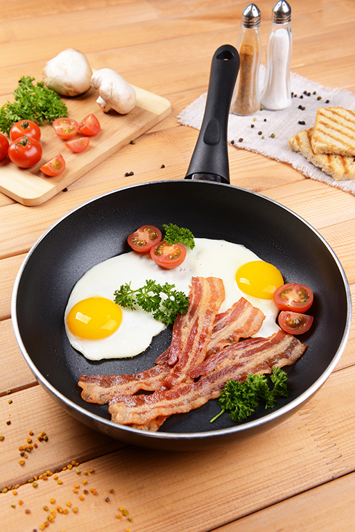 A ceramic pan with a fried egg and bacon on a wooden table, spices and vegetables in the background.